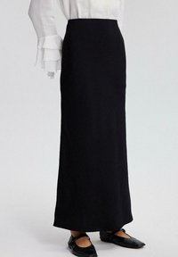Falda negra hasta los tobillos, hecha de una tela suave, con una cintura ajustada y una silueta recta, combinada con zapatos planos negros y una blusa blanca.