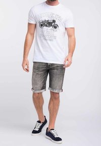 Camiseta blanca con estampado gráfico negro, pantalones cortos de mezclilla gris con dobladillo enrolado y zapatillas oscuras con cordones blancos.
