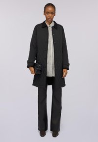 Černý prošívaný kabát s límečkem, předními knoflíky a bočními kapsami, spárovaný se pruhovanou košilí s knoflíky a tmavými kalhotami.