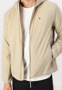 Mężczyzna noszący beżową kurtkę zapinaną na zamek na białą koszulkę i czarne spodnie, z ręką w prawej kieszeni, widoczny tors bez głowy i stóp.