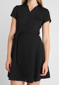 Vestido negro de manga corta con escote en V abotonado, cintura ceñida y cinturón de lazo. Fabricado con una tela ligera y fluida, con una falda hasta la rodilla.