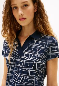 Μπλε ναυτικό πόλο μπλουζάκι με λευκό αφηρημένο μοτίβο. Κοντά μανίκια, επίπεδη γιακάς και μικρή λεπτομέρεια λογότυπου στο στήθος. Υλικό από βαμβακερό μείγμα.