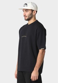 Black cotton t-shirt with short sleeves; features gold text "PAS DE MONDE" on the chest; worn with a light-colored cap.