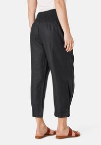 Zwarte linnen cropped broek met een smocktail, relaxte pasvorm en knoopdetail aan de zoom, gecombineerd met bruine schuifsandalen.