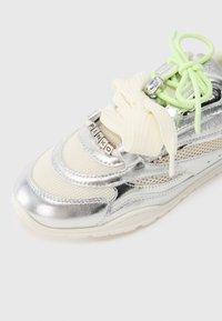 Ασημί και κρεμ αθλητικό παπούτσι με ύφασμα από δίχτυ, μεταλλικές λεπτομέρειες, κορδόνια νέον πράσινου χρώματος και διακοσμητικό φιόγκο. Λογότυπο στην πλευρά.