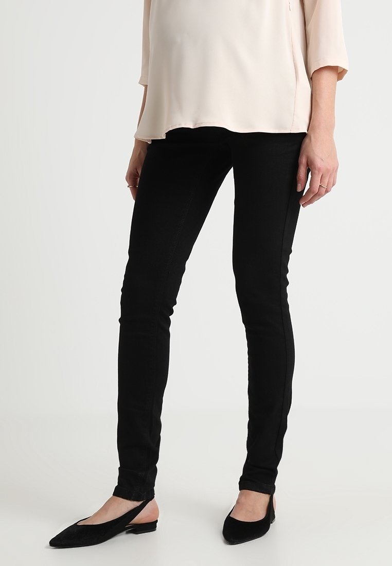 Femme portant un pantalon noir slim, un chemisier beige et des ballerines noires à bride arrière, debout devant un fond clair uni.