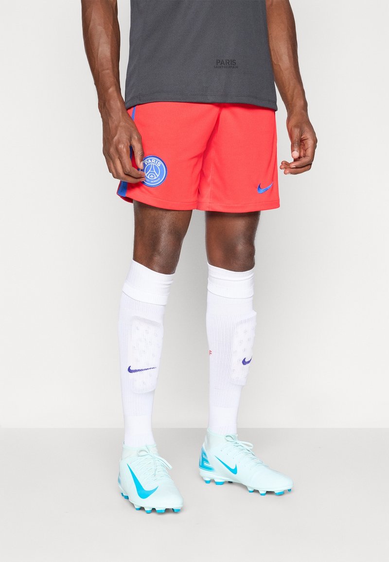 Joueur de football masculin portant un short rouge du Paris Saint-Germain, des chaussettes blanches avec des protège-genoux et des crampons Nike bleu clair, debout devant un fond blanc.