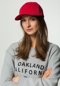 Red baseball cap with a flat brim, mesh side panels, and a structured design, worn with a gray sweater featuring black lettering.