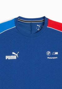 Zila T-kreklus ar sarkaniem un gaiši ziliem piedurkņu, kurā ir balti logo: Puma priekšpusē un BMW Motorsport sānos.