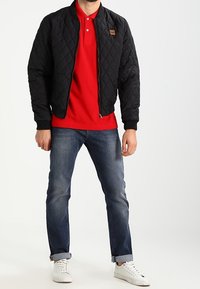 Černá prošívaná bomber bunda, červené polo tričko, modré džíny a bílé tenisky. Viditelné logo na bundě, džíny s ohrnutými nohavicemi.