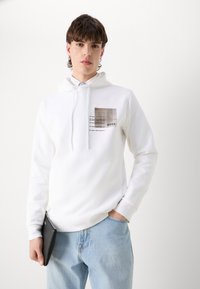Sweat à capuche blanc en tissu mélangé de coton, doté d'un patch texturé avec un motif imprimé, de coutures contrastées et d'une poche kangourou.