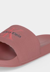 Ροζ σανδάλι Calvin Klein slide με υφασμάτινο λουράκι που έχει ανάγλυφο κόκκινο «CK» και μαύρο κείμενο «Calvin Klein».