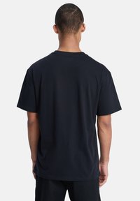 Homme de dos faisant face à l'appareil photo, portant un T-shirt noir uni à manches courtes et un pantalon noir, sur fond blanc.