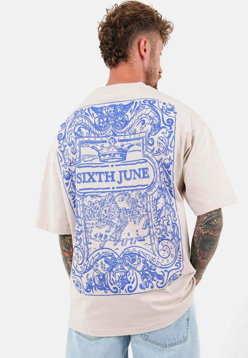 Camiseta de gran tamaño en color beige con un gran diseño gráfico en azul en la parte posterior, que presenta patrones ornamentales y el texto "SIXTH JUNE."