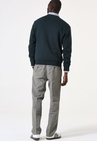 Sudadera verde oscura con puños de canalé, combinada con pantalones gris claro. Un notable cuello de camisa con patrón blanco emerge de debajo. Zapatillas blancas.