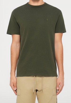 Homme portant un t-shirt vert olive à manches courtes avec petit logo brodé et pantalon cargo beige avec taille à cordon.