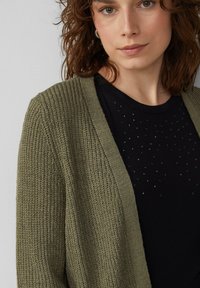 Gilet en tricot vert olive avec un motif texturé, à ouverture devant et bords côtelés, porté sur un haut noir orné de petits points décoratifs.