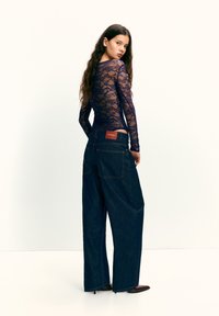 Granatowa koronkowa bluzka z długim rękawem w połączeniu z luźnymi, ciemnymi dżinsowymi spodniami z dużymi kieszeniami i czerwoną metką na pasku.