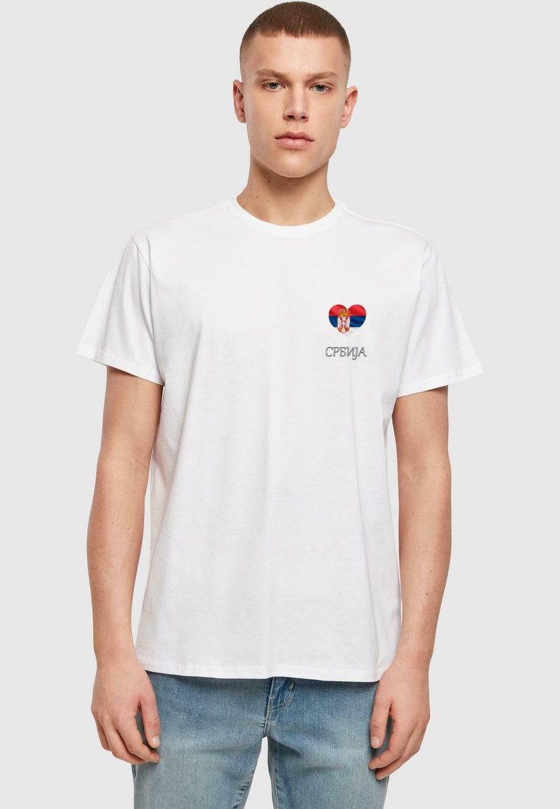 Hvit bomull t-skjorte med et rødt og blått hjerte-design med et serbisk emblem og teksten "СРБИЈА" i grå. Klassisk passform, korte ermer.