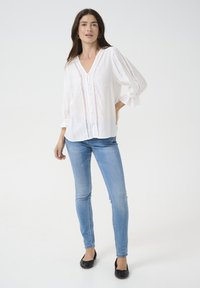 White cotton blouse with a V-neck and button closure, featuring lace detailing along the front and cuffs. Paired with light blue skinny jeans.
