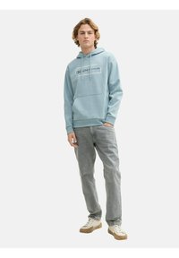 Sudadera azul claro con bolsillo frontal, capucha con cordones y logotipo impreso. Combinada con jeans gris claro y zapatillas beige. Textura de tela suave.