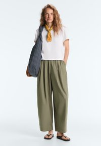 Frau trägt weißes T-Shirt, olivgrüne weit geschnittene Hose, senffarbener Halsschal, schwarze Flip-Flops und hält eine gestreifte Umhängetasche vor weißem Hintergrund.