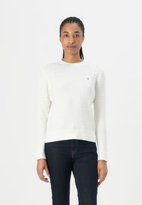 Kvinne med flettet hår som har på seg en hvit teksturert genser og mørke jeans, stående mot en enkel hvit bakgrunn.