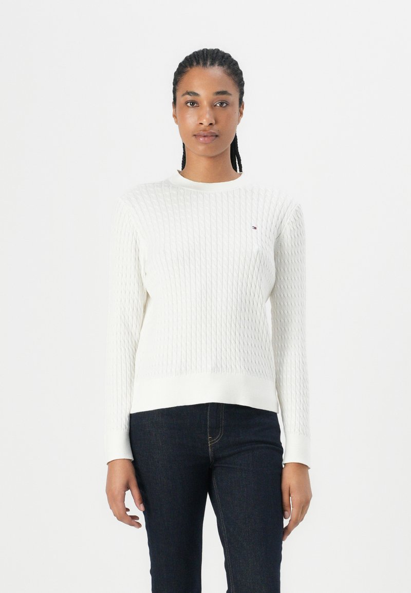 Kvinne med flettet hår som har på seg en hvit teksturert genser og mørke jeans, stående mot en enkel hvit bakgrunn.