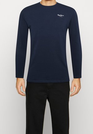 Mies, joka pukeutunut tummansiniseen pitkähihainen pyöreäkauluspaitaan, jossa on valkoinen "Pepe Jeans London" -logo rinnassa, ja mustiin housuihin.