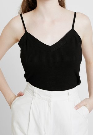 Kobieta ubrana w czarny bezrękawnik prążkowany z cienkimi ramiączkami, wpuszczony w wysokie białe spodnie, z rękami w kieszeniach.