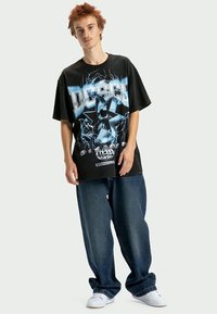 Jauns vīrietis ar adatu sarkaniem matiem, ģērbies melnā pārspīlētā grafiskajā t-kreklā, brīvās tumši zilās džinsās un baltās sporta apavās, stāv pretī vienkāršai fona.