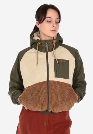 Frau trägt eine farbblockierte Kapuzenjacke mit olivgrünen Ärmeln, cremefarbenem Oberteil und brauner Fleece-Unterseite, Hände in den Taschen.