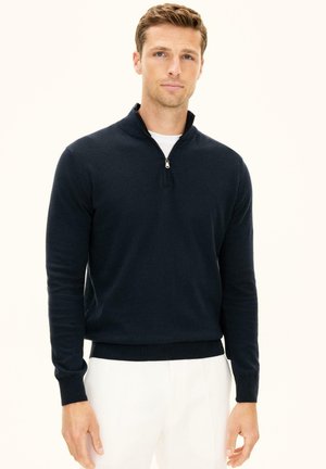 Giovane uomo con capelli corti che indossa un maglione blu navy con cerniera a quarto sopra una camicia bianca e pantaloni bianchi, in piedi davanti a uno sfondo semplice.