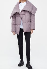 Puffjakke i lys lavendel med en unik oversized krave, der har en quiltet design, trykknaplukninger og en afslappet pasform. Kombineret med sorte bukser og støvler.
