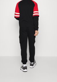 Person, der schwarze Cargo-Jogginghosen, einen schwarz-roten Sweatshirt mit weißen Streifen an den Ärmeln und graue Sneakers trägt, steht vor einem weißen Hintergrund.
