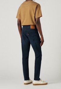 Mörkblå denimjeans med rak passform, i kombination med en brun t-shirt. Funktioner inkluderar bakfickor och en läderlapp vid midjan.