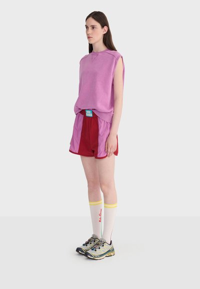 Jeune femme debout de profil portant un haut violet sans manches, un short rouge et violet, des chaussettes blanches montantes avec des rayures jaunes, et des baskets grises.