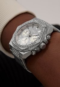 Montre chronographe octogonale argentée avec un cadran texturé, deux sous-cadrans et des accents en cristal sur la lunette et le bracelet. Portée au poignet.