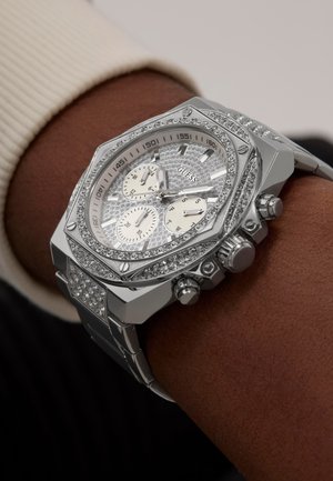 Montre chronographe octogonale argentée avec un cadran texturé, deux sous-cadrans et des accents en cristal sur la lunette et le bracelet. Portée au poignet.
