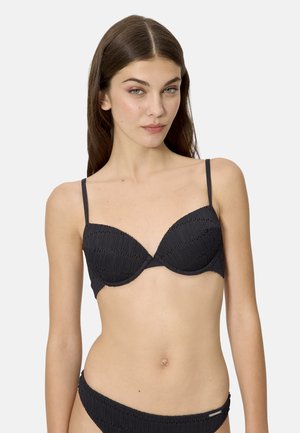 Mujer con bikini negro texturizado de tiras delgadas, de pie frente a un fondo liso de color gris claro, mirando hacia adelante.