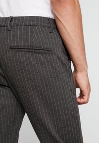 Szare spodnie w prążki wykonane z tkaniny o gładkiej fakturze. Posiadają boczne kieszenie oraz tylną kieszeń z obszyciem.