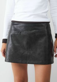 Schwarzer Leder-Minirock mit zwei vorderen Taschen, glatter Textur und geradem Schnitt. Kombiniert mit einem weißen Langarm-Oberteil.