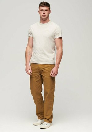 Hellgraues Baumwoll-T-Shirt und braune Hose mit Seitentaschen. Das T-Shirt hat ein kleines Logo; die Hose hat eine Steppnaht und ist gerade geschnitten.