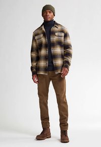 Kariertes Flanellhemd in Beige und Navy mit brauner Hose; trägt einen schwarzen Rollkragenpullover und eine olivgrüne Mütze; braune Wanderschuhe.