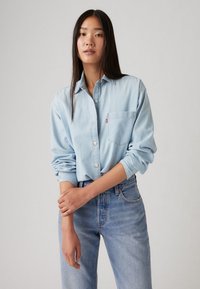 Camicia di jeans azzurri con apertura a bottoni e taschino sul petto, abbinata a jeans azzurri a vita alta, che mostrano una vestibilità rilassata.