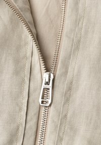 Gros plan sur un tissu beige avec une fermeture éclair métallique argentée partiellement ouverte, étiquetée "STREET ONE" sur un vêtement.