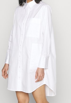 Donna che indossa un vestito camicia bianco lungo, a maniche lunghe e dal taglio morbido, con colletto e tasca sul petto, in piedi contro uno sfondo neutro.