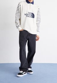 Poloko s dlouhým rukávem se proužkovanými rukávy, krémovým tělem, námořnickým límcem a logem. Spárováno s tmavě šedými džíny a černo-bílými teniskami.