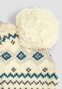 Strikket hue i cremefarve med teal og mørkeblå diamantmønstre. Har en stor, fluffy hvid pom-pom på toppen. Blød tekstur.
