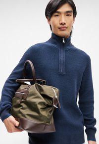 Oliven grøn taske med brune læderdetaljer og lynlåslommer. Vævet mørkeblå sweater med høj krave. Glat tekstur, struktureret form.